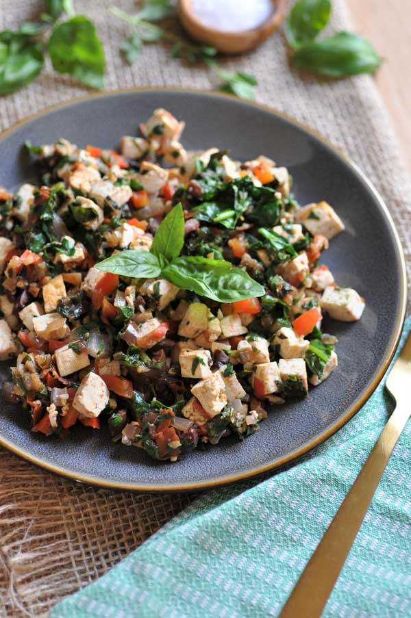 Mediterranean tofu scramble on a grey plate with a green napkin and gold fork next to it and fresh herbs sprinkled around it. 