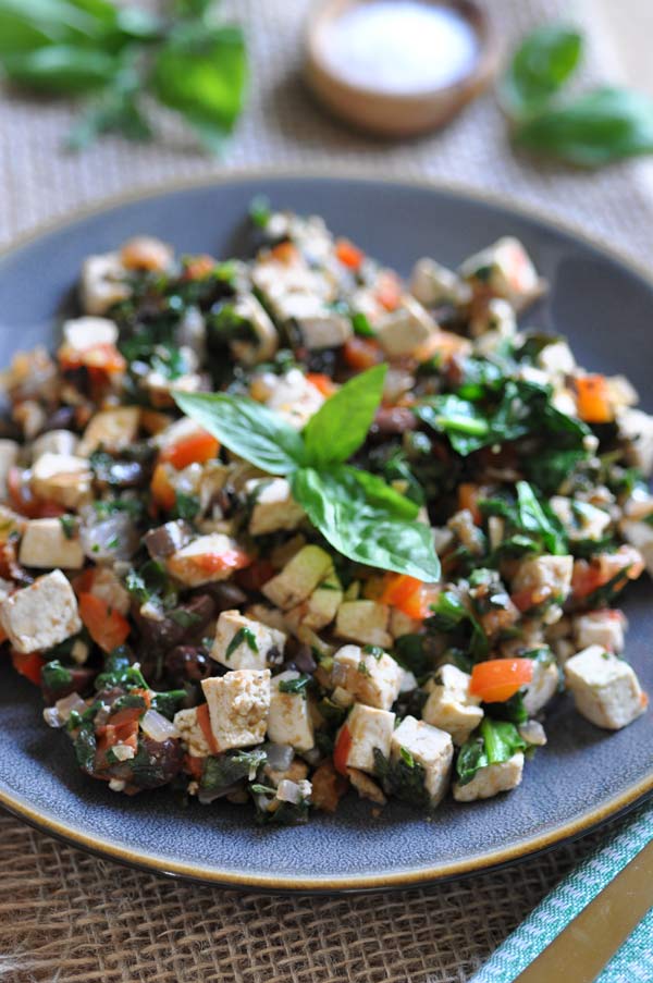 Close up of Mediterranean Tofu Scramble! on a grey plate with a sprig of basil in the center and a small wood bowl with sea salt and fresh herbs int he background