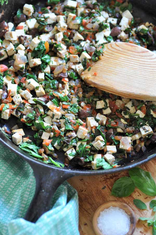 Mediterranean Tofu Scramble in an iron skillet with a wood spoon in the pan and a green towel wrapped around the handle.