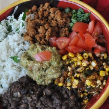 A burrito bowl with black beans, rice, corn, tomatoes, and gaucamole, and a silver fork next to the bowl.