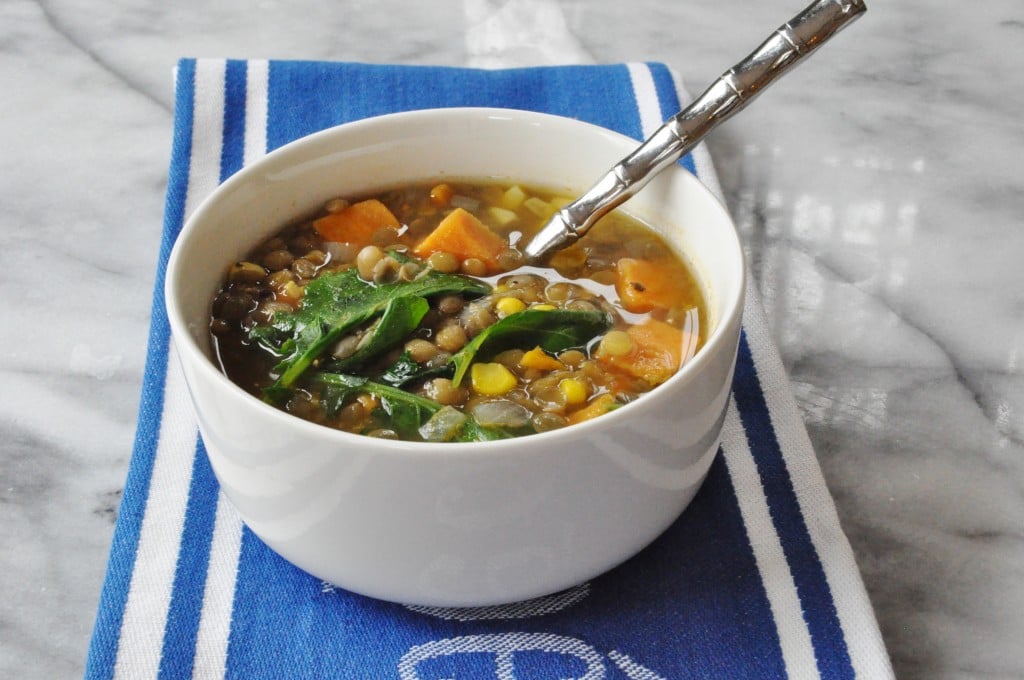 A white bowl filled with vegetable soup with a silver spoon in it on a blue and white towel on a marble bo