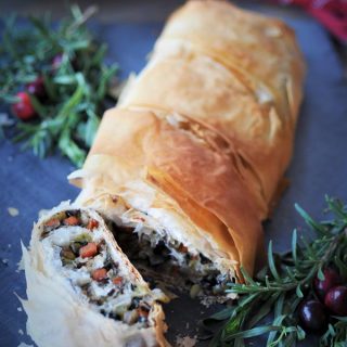 Sliced Vegan Vegetable Wellington on a gray piece of slate with fresh sprigs of tarragon, rosemary, and a few fresh cranberries for decoration.