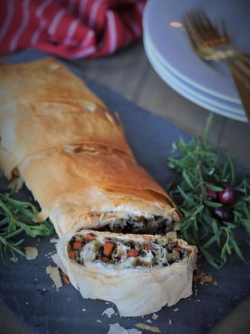 A sliced vegetable wellington on a gray slate board on a wooden table with fresh herbs for garnish and white plates, gold forks, and a red towel in the background.