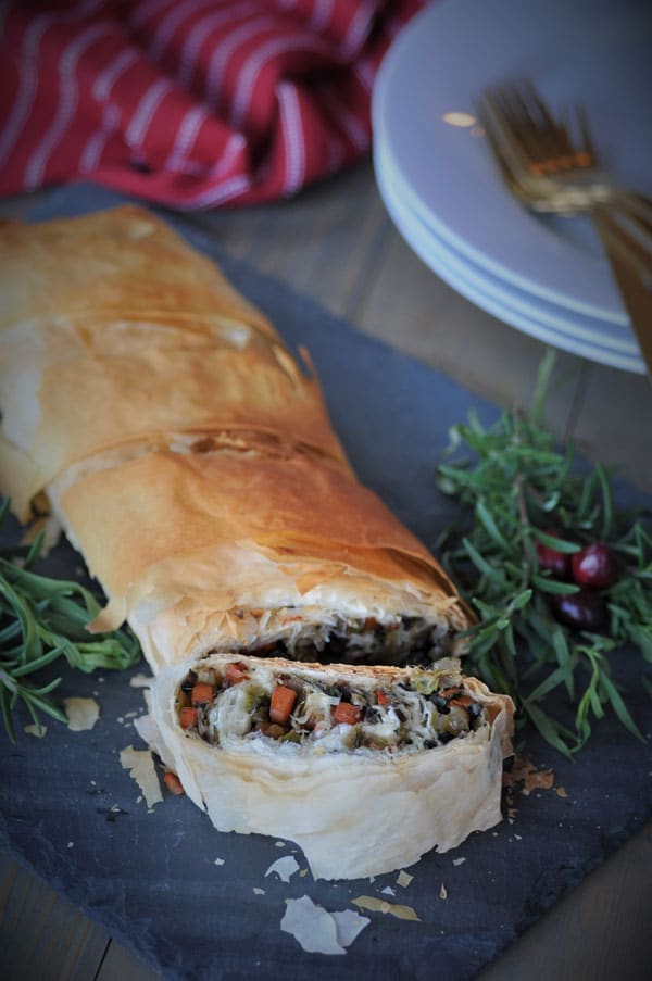 A sliced vegetable wellington on a gray slate board on a wooden table with fresh herbs for garnish and white plates, gold forks, and a red towel in the background.