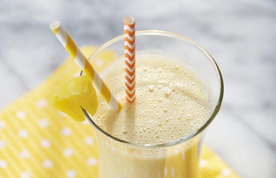 A glass filled with a golden smoothie with a slice of pineapple and an orange and white straw.