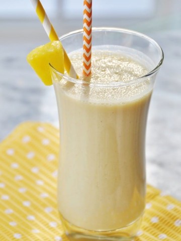 A glass with a yellow smoothie with a pineapple wedge on the edge of the glass and an orange and white straw on a yellow and white polka dot napkin.