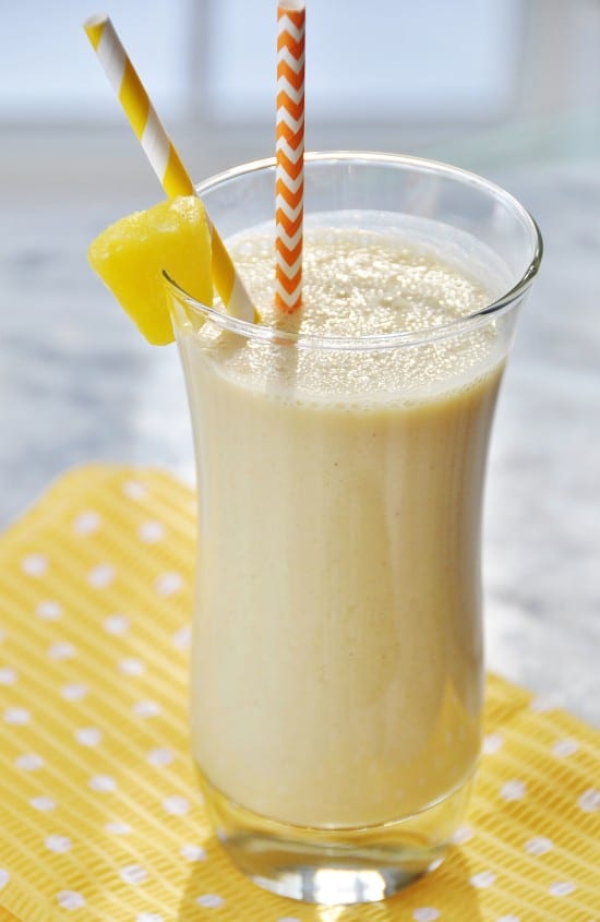 A glass with a yellow smoothie with a pineapple wedge on the edge of the glass and an orange and white straw on a yellow and white polka dot napkin.