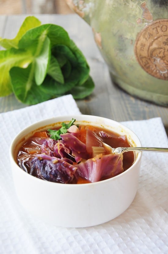 Sopa de col roja en un bol blanco con una cuchara de plata y verduras al fondo.