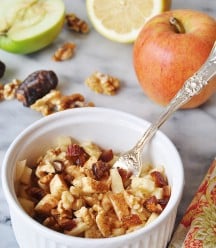 Raw Apple Pie in a white bowl with a silver fork in the bowl, and a red apple and half of a green apple and a lemon, and walnuts in the background