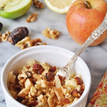 Raw Apple Pie in a white bowl with a silver fork in the bowl, and a red apple and half of a green apple and a lemon, and walnuts in the background