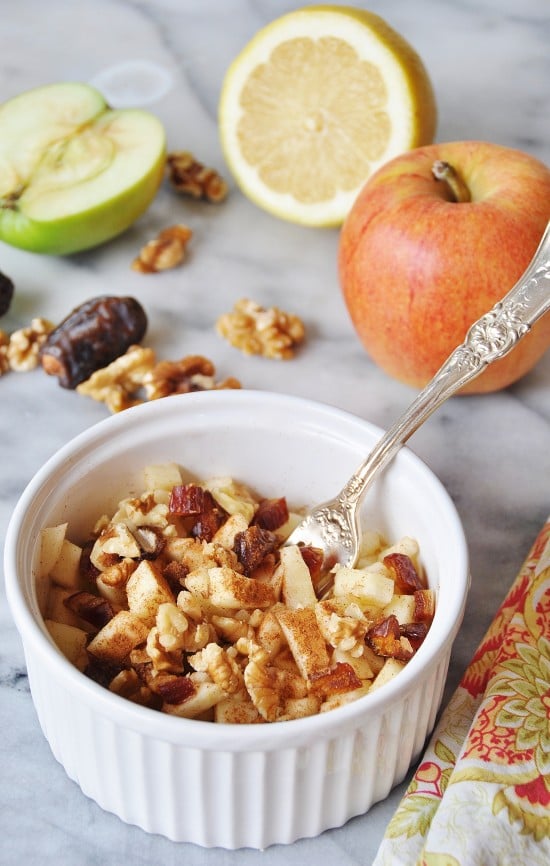 Raw Apple Pie in a white bowl with a silver fork in the bowl, and a red apple and half of a green apple and a lemon, and walnuts in the background