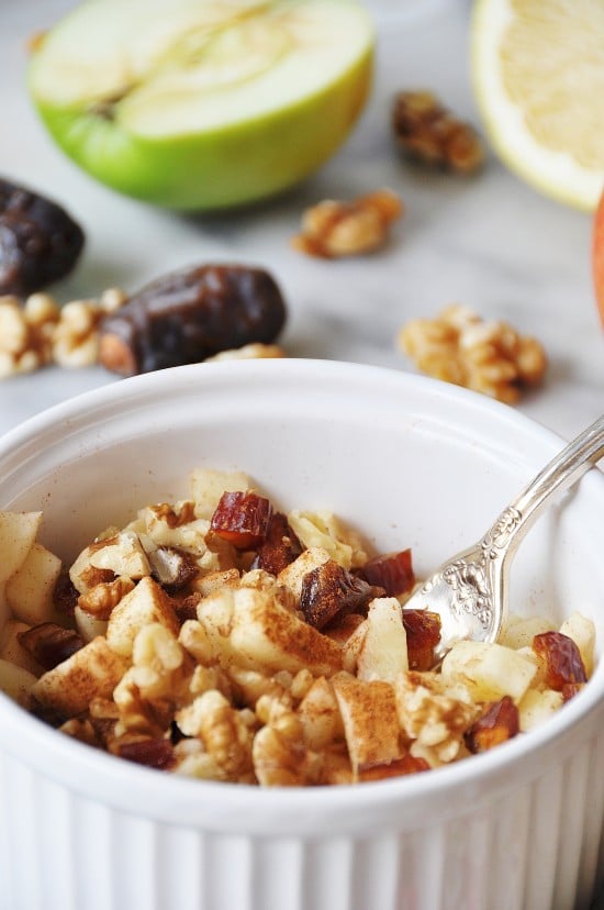 Raw Apple Pie in a white bowl with a silver fork and dates, apples, and walnuts next to it. 