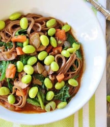 A white bowl with soba noodles, broth, vegetables, and edamame on a green and white towel.