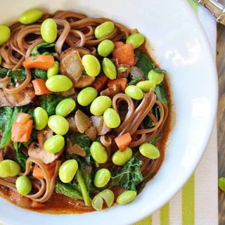 A white bowl with soba noodles, broth, vegetables, and edamame on a green and white towel.