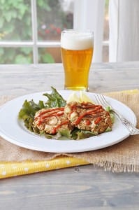 Crab cakes on a bed of lettuce on a white plate with sriracha drizzled on top with a beer in the background.