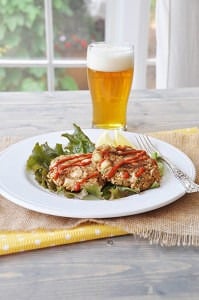 Crab cakes on a bed of lettuce on a white plate with sriracha drizzled on top with a beer in the background.