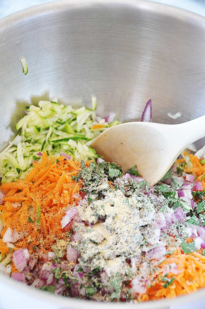 A silver mixing bowl with shredded zucchini, carrots, chopped red onion, breadcrumbs, and spices being stirred with a wood spoon.