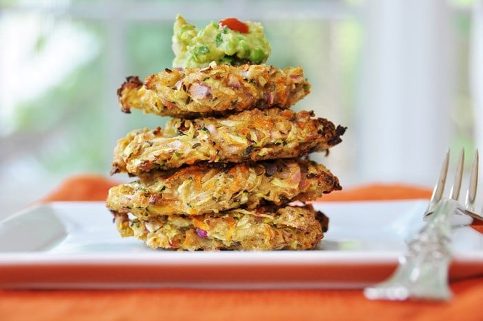 Four Vegan Oil-Free Zucchini & Carrot Fritters stacked on a square white plate with a dollop of guacamole on top and a silver fork. 