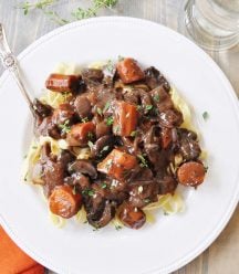 Mushroom Fourguignonne on a white plate with a silver fork and an orange napkin tucked under the bottom left corner of the plate.