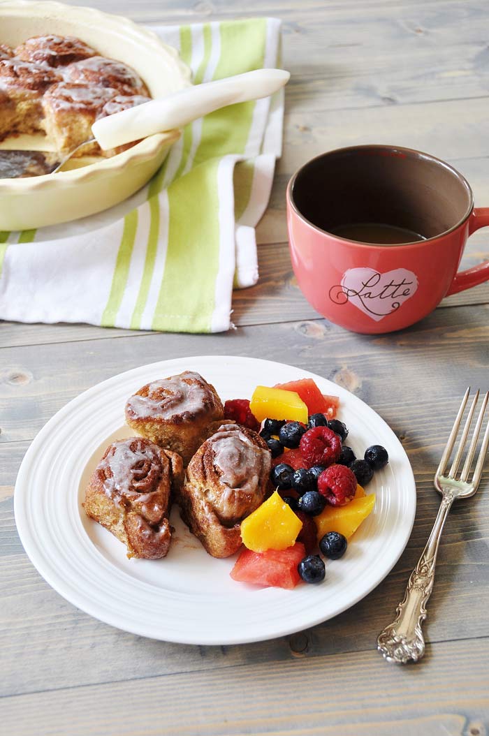 Homemade Vegan Cinnamon Rolls and fruit salad on a white plate with a pink mug of coffee and a pan of rolls next to it