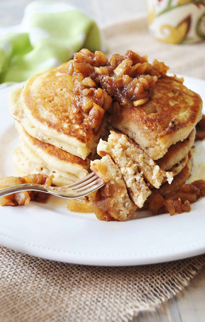 A stack of 3 pancakes with maple syrup and chunky applesauce on a white plate with a silver forkful of the pancakes.