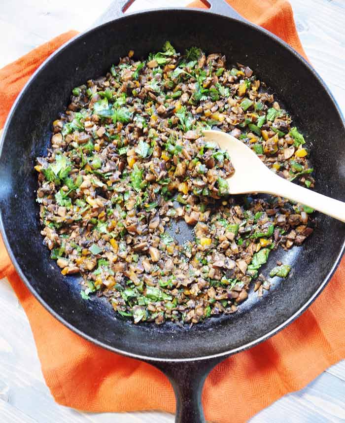 A cast-iron skillet filled with mushroom tinga on an orange towel with a wood spoon scooping up the tinga.