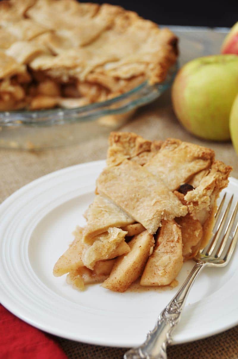 A slice of Homemade Apple Cinnamon Pie with a Flaky Vegan Pie Crust! on a white plate with a silver fork and the whole pie and an apple in the background