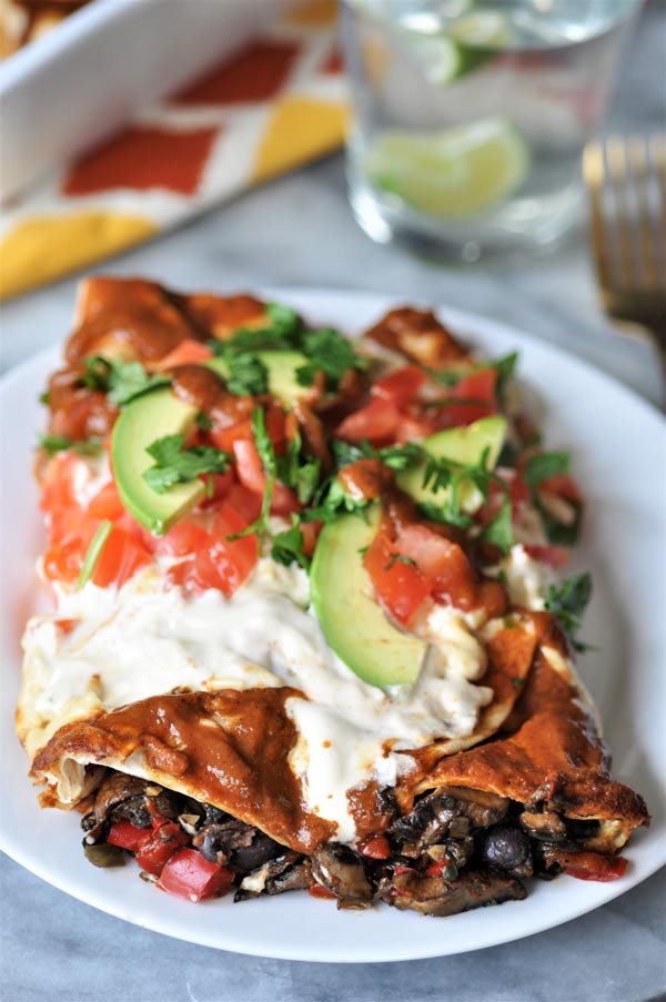 Two black bean and mushroom enchiladas on a white plate, garnished with chopped cilantro, tomato, and sliced avocado.