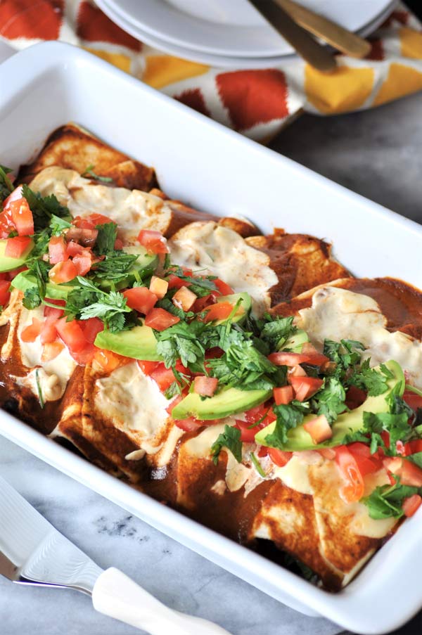 A white pan full of vegan black bean & mushroom enchiladas with sliced avocado, cilantro, and chopped tomato on top.