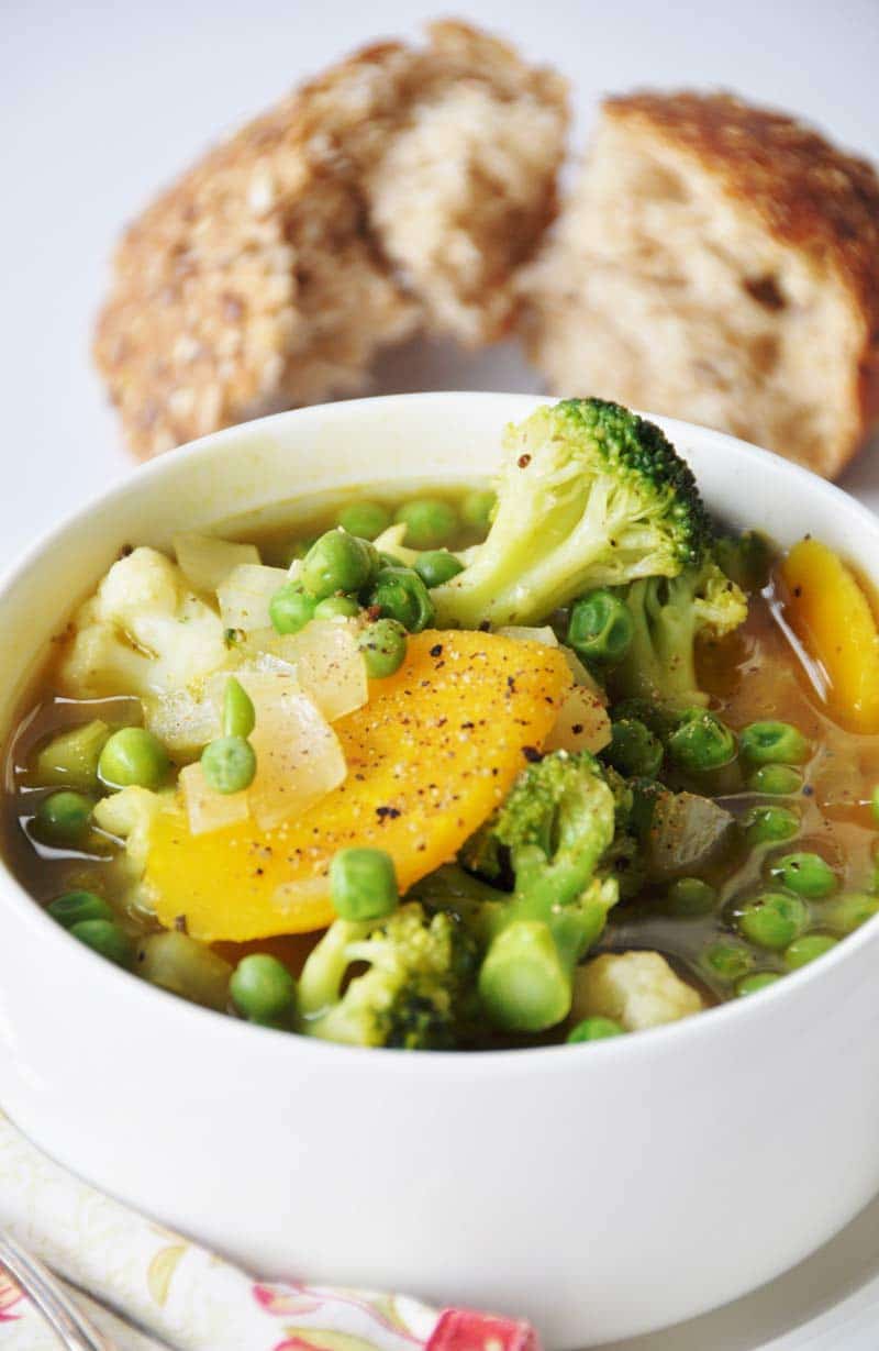 A close up picture of 7 Ingredient 30 Minute Vegetable Soup in a white bowl, sprinkled with black pepper, with torn wheat bread behind the bowl and the handle of a silver spoon on a red and yellow napkin tucked next to the bowl