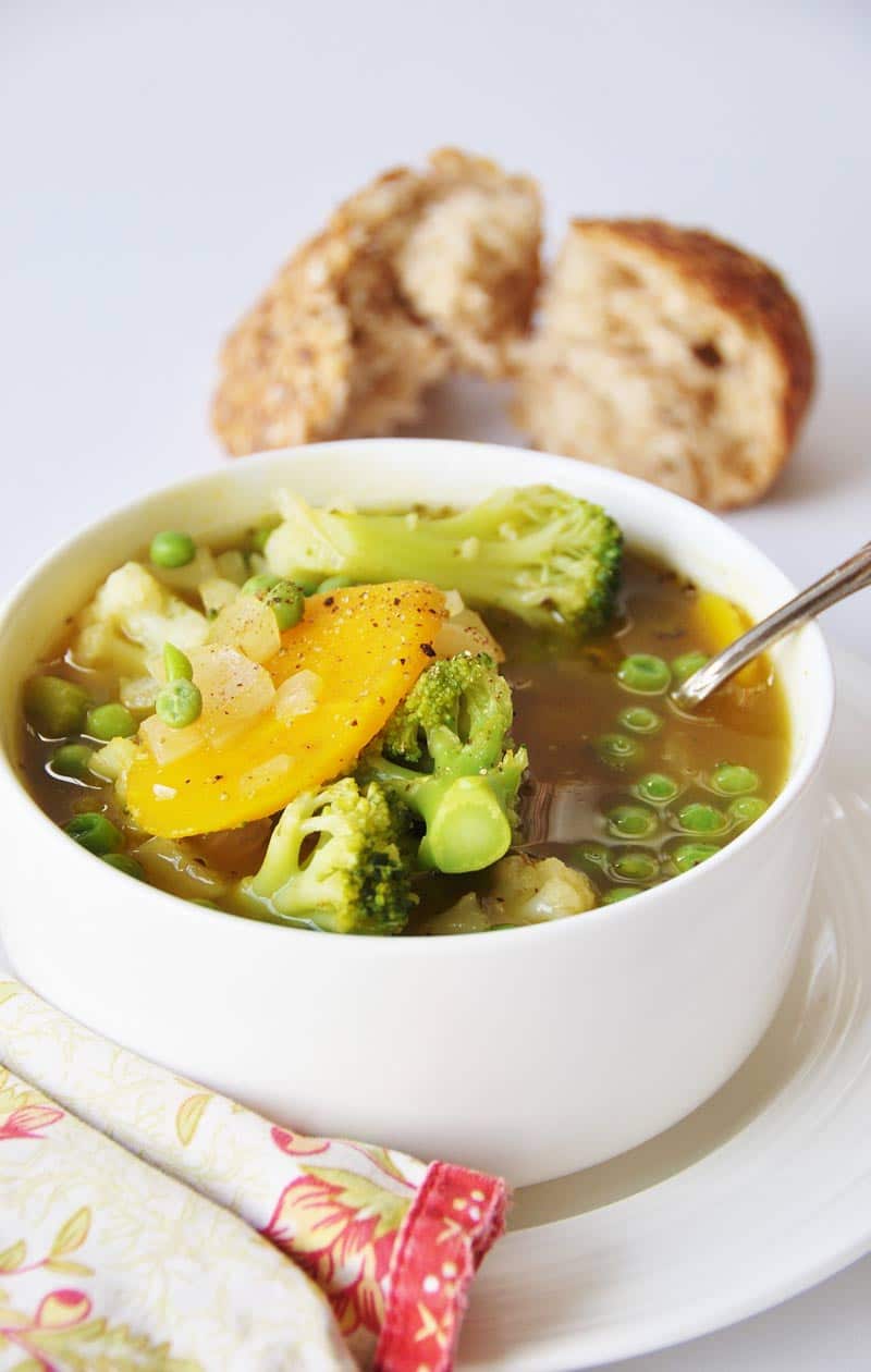 7 Ingredient 30 Minute Vegetable Soup in a white bowl on a white plate with a silver spoon sticking out of the bowl and a red and yellow floral napkin on the edge of the plate and pieces of torn bread in the background