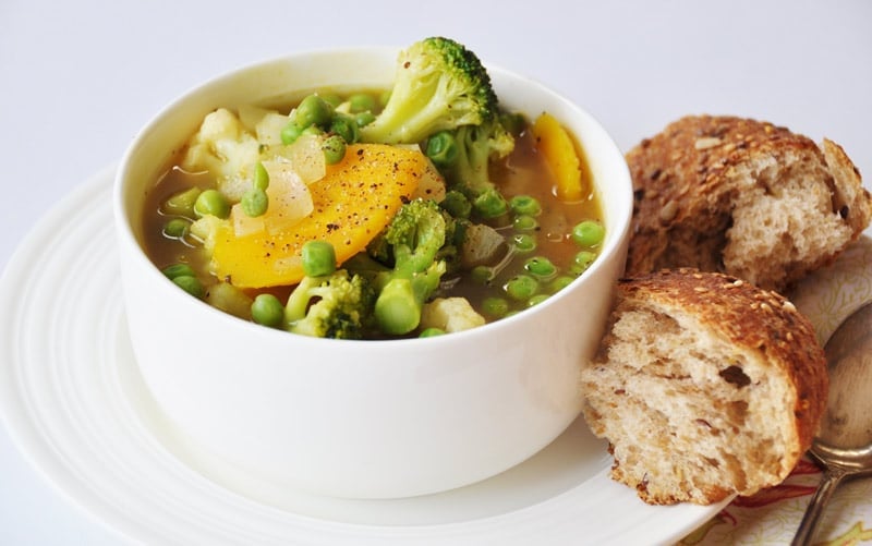 7 Ingredient 30 Minute Vegetable Soup in a white bowl on a white plate with two pieces of grain bread on the side of the plate and a silver spoon on a yellow and red floral napkin next to it.