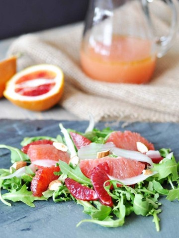 A salad with greens and sliced citrus on a slate board with a glass pitcher of orange dressing in the background with a sliced grapefruit.