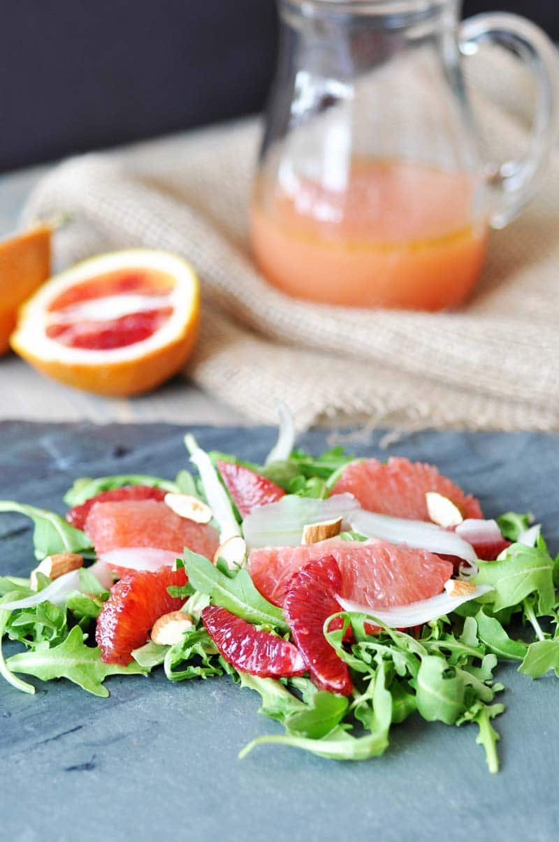 A salad with greens and sliced citrus on a slate board with a glass pitcher of orange dressing in the background with a sliced grapefruit.