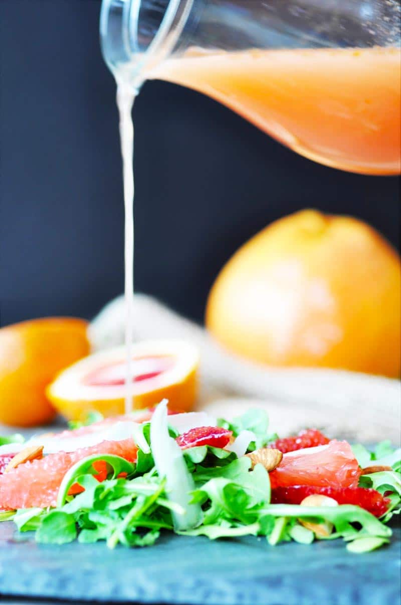 A pitcher of orange salad dressing being poured on a salad with sliced citrus and arugula. 