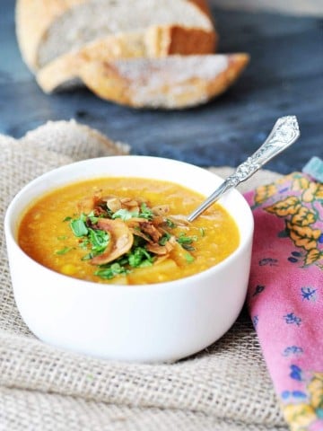 Corn chowder in a white bowl with a silver spoon sticking out of it and a loaf of sliced bread in the background.