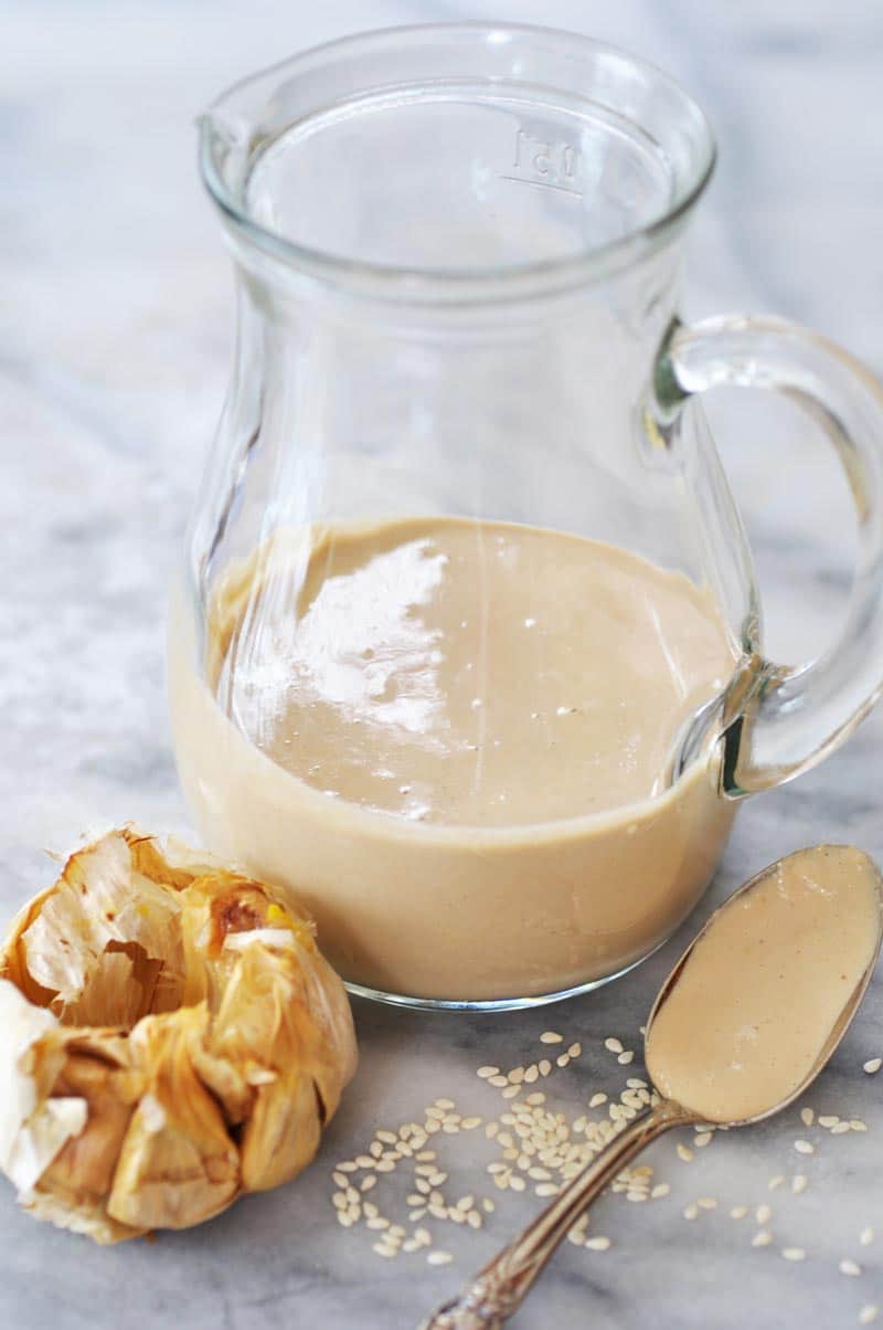 A glass pitcher with roasted garlic tahini dressing in it and a spoon with the dressing on it and a bulb of roasted garlic and sesame seeds next to it.