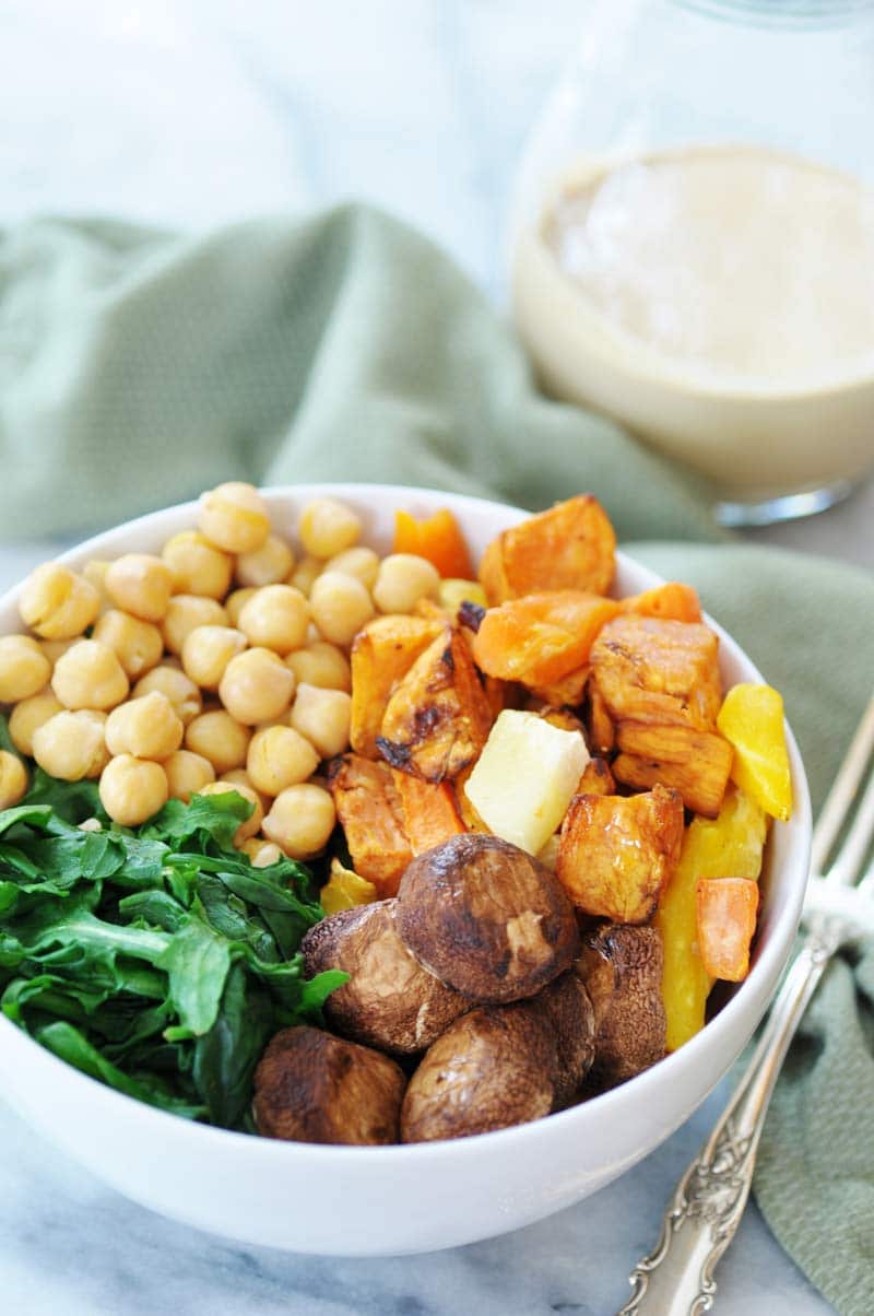 Roasted mushroom caps, sweet potatoes, garbanzo beans, and spinach in a white bowl with a silver fork, green napkin, and pitcher of tahini dressing next to it.