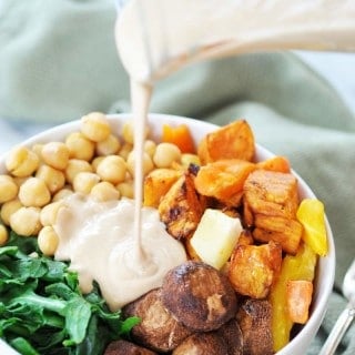 A white bowl filled with roasted vegetables with a glass pitcher of dressing being poured over it