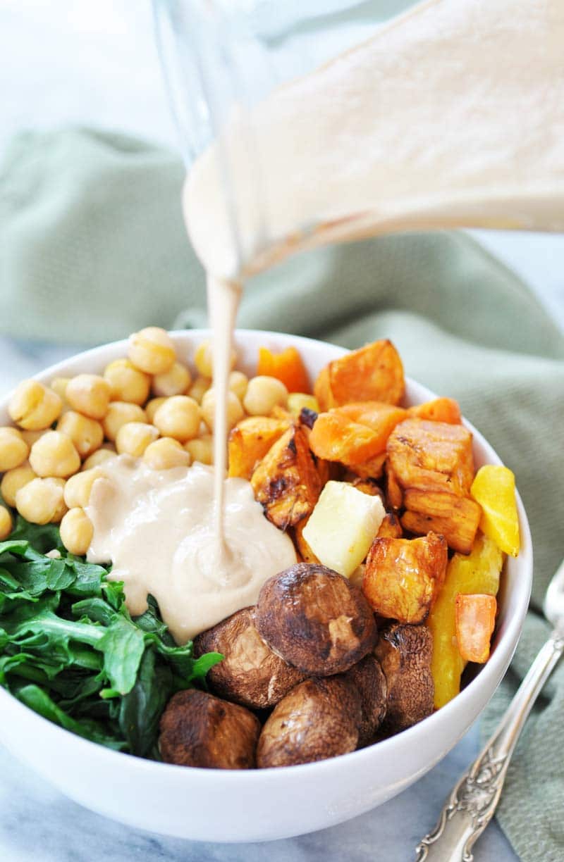 Roasted mushrooms, sweet potatoes, spinach, and chickpeas in a white bowl with garlic tahini dressing being poured on top of it.