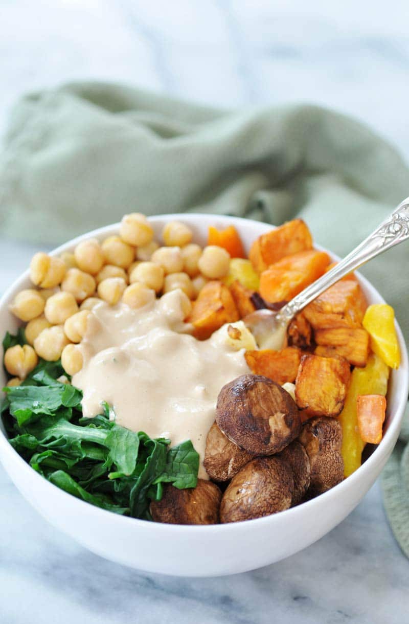 Roasted mushroom caps and sweet potatoes with spinach and chickpeas, and garlic tahini dressing in the center with a silver fork in the bowl