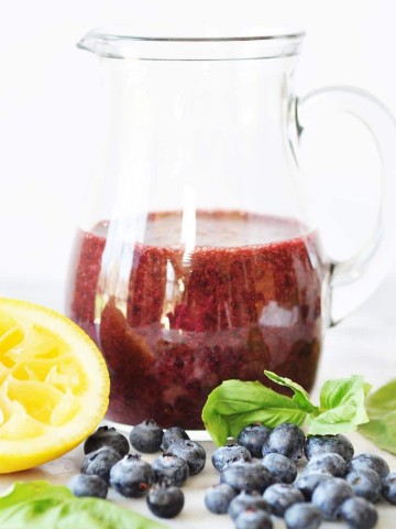 Blueberry Basil Salad Dressing in a clear glass pitcher with a lemon slice, fresh blueberries, and basil around it on a white surface
