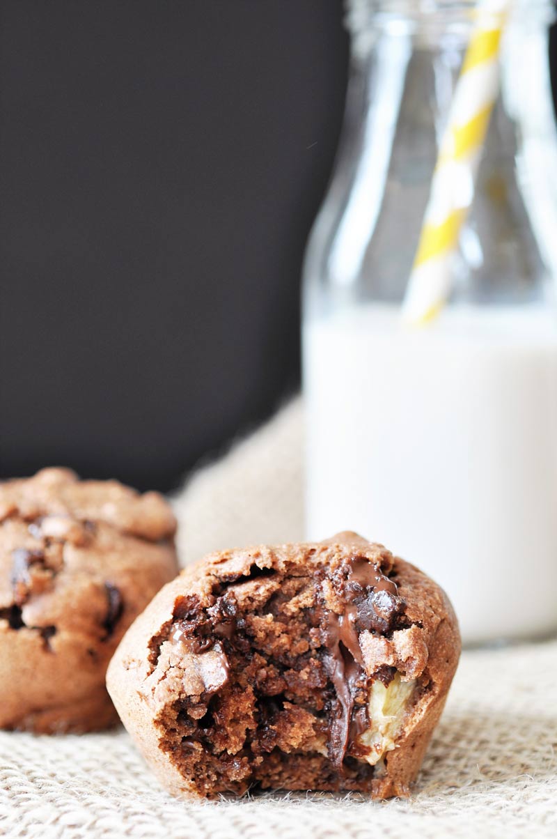 A chocolate banana muffin with a bite taken out of it and a bottle of milk with a yellow and white straw in it and more muffins in the background. 