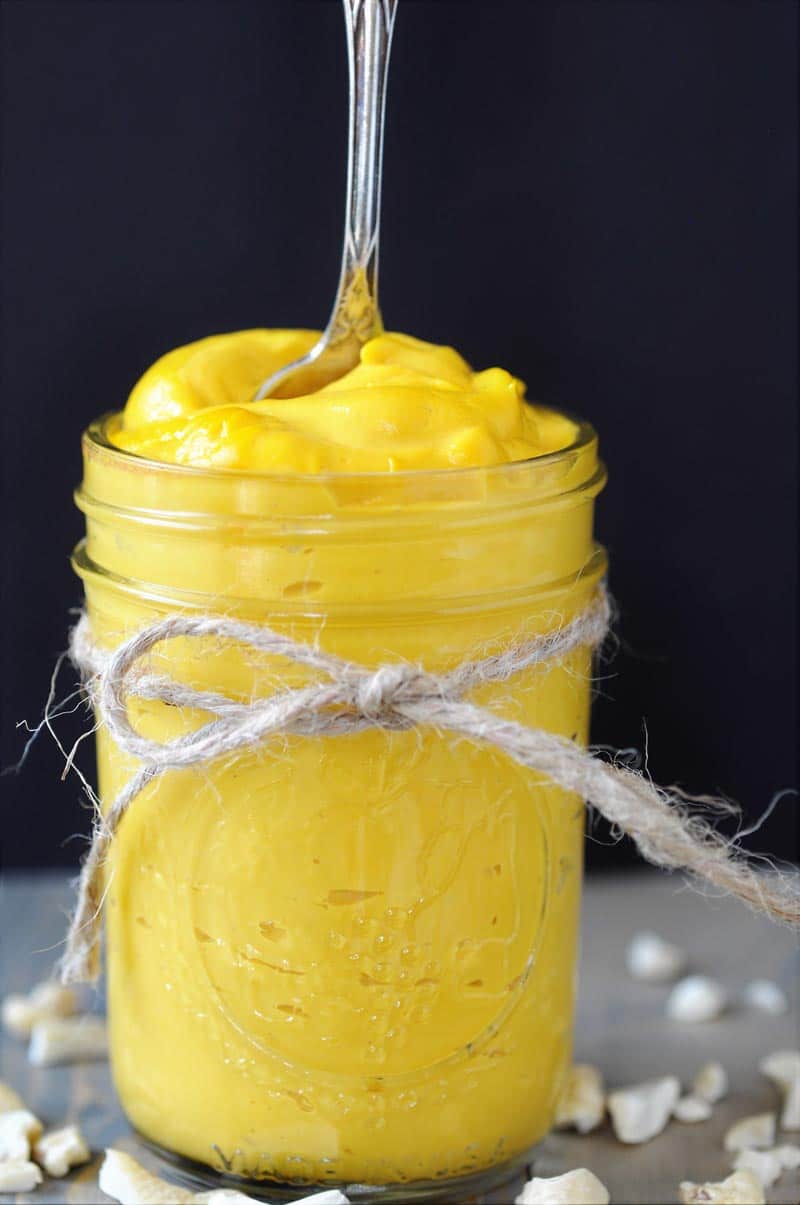 A cheddar cheese sauce in a mason jar with a piece of rope tied around the jar and a silver spoon standing in the sauce, and cashews spread around the jar.