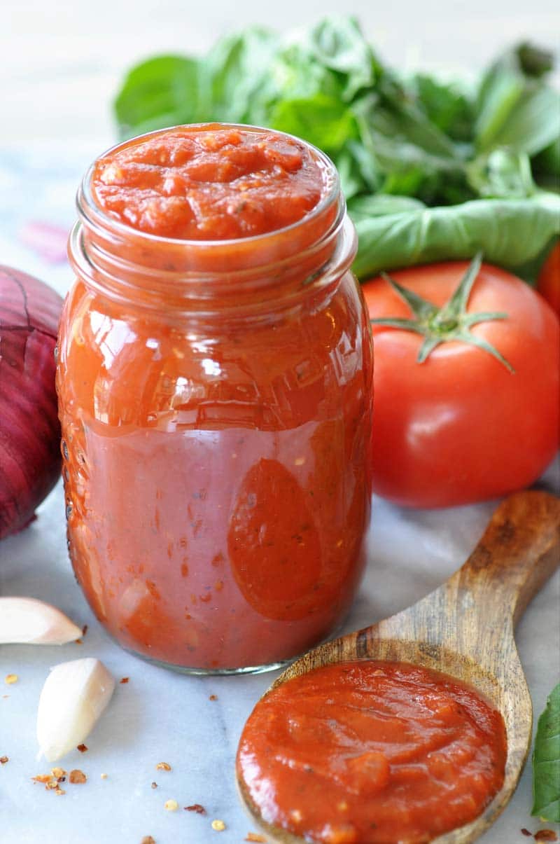 A mason jar filled with pizza sauce and a wooden spoon with the sauce in it in the front of the jar and tomatoes, basil and garlic around the jar.