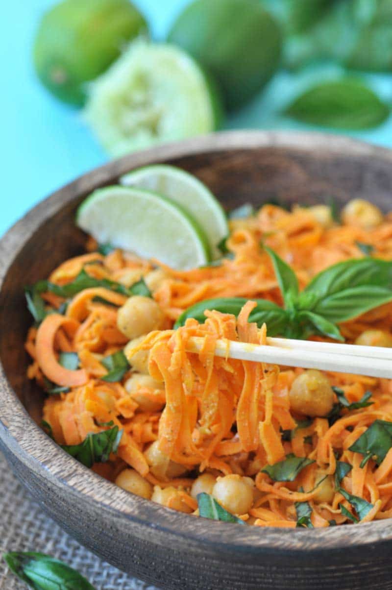 A pair of chopsticks lifting sweet potato noodles out of a wooden bowl that's filled with the noodles, chickpeas, basil, and two lime wedges.