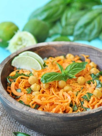 A wooden bowl with sweet potato noodles, chickpeas, and basil and lime wedges, with a pile of basil and limes in the background.