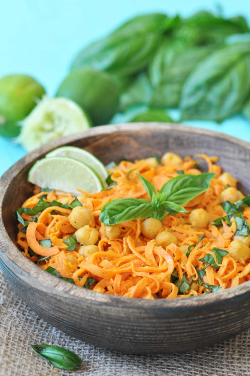 A wooden bowl with sweet potato noodles, chickpeas, and basil and lime wedges, with a pile of basil and limes in the background. 