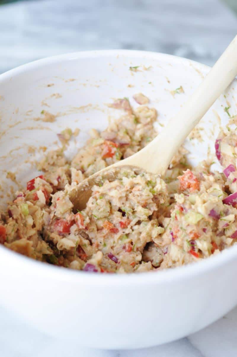 A wooden spoon stirring tuna salad in a white bowl.