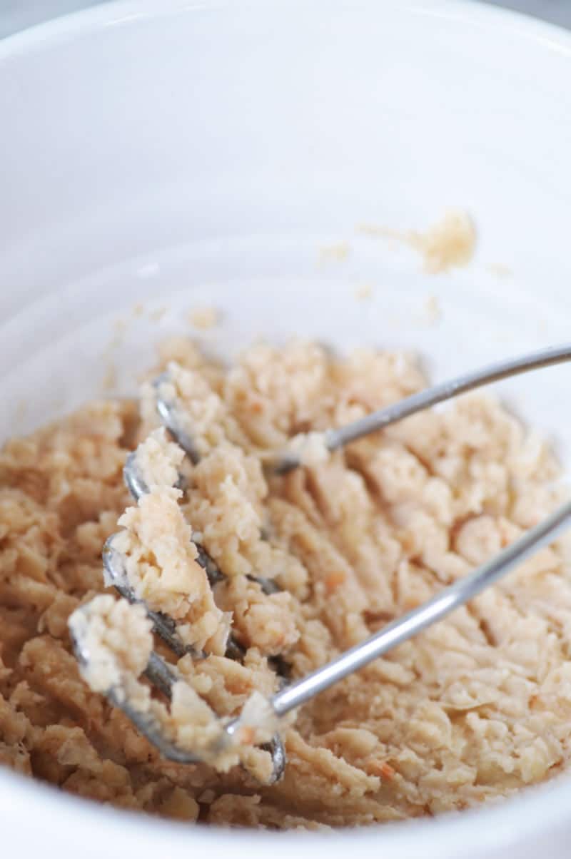 A potato masher that's mashing white beans in a white bowl.